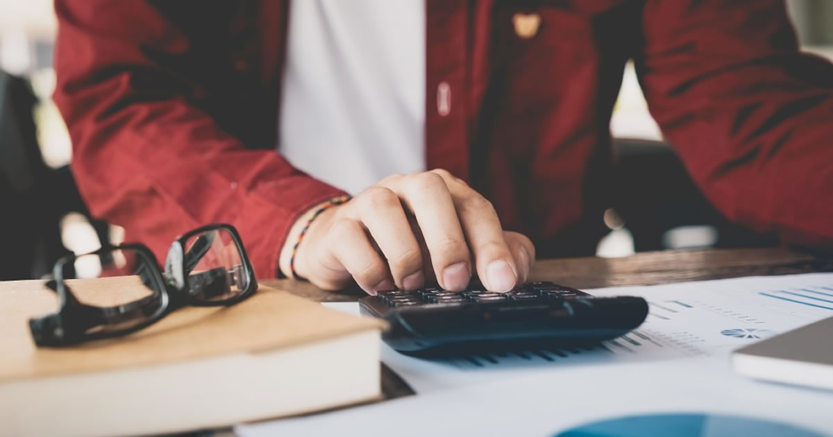 Close up of hand using calculator on desk with book, eyeglasses, and papers