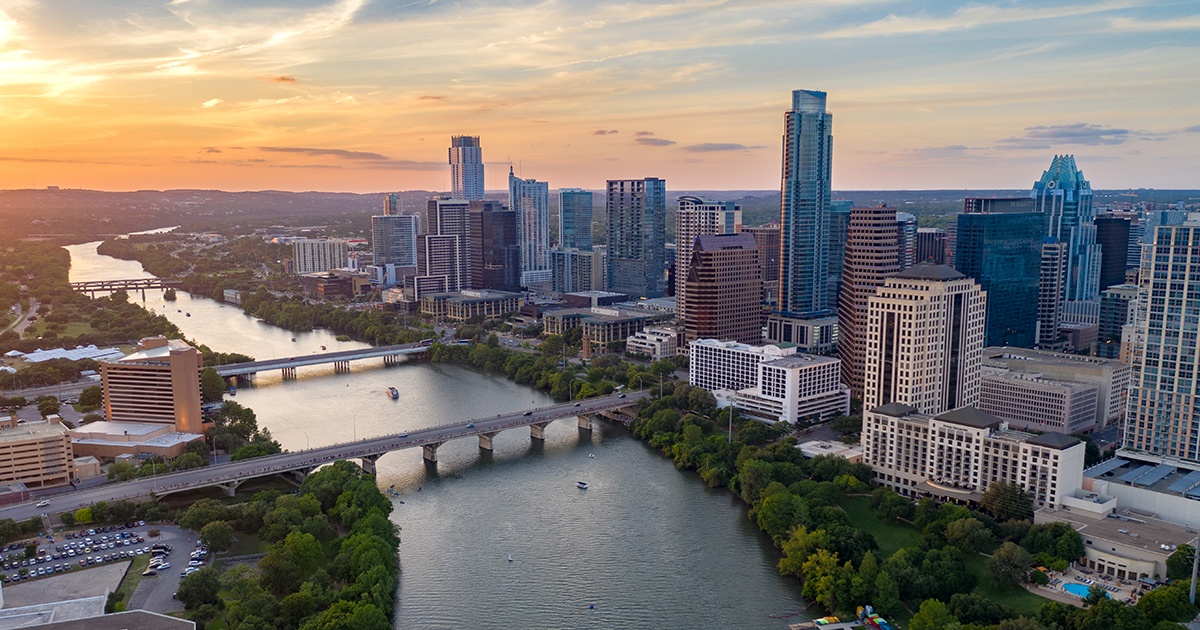 Aerial view of downtown Austin, Texas at sunset, featuring the Colorado River winding through the city, multiple bridges crossing the water, and a skyline of tall modern buildings surrounded by green spaces.